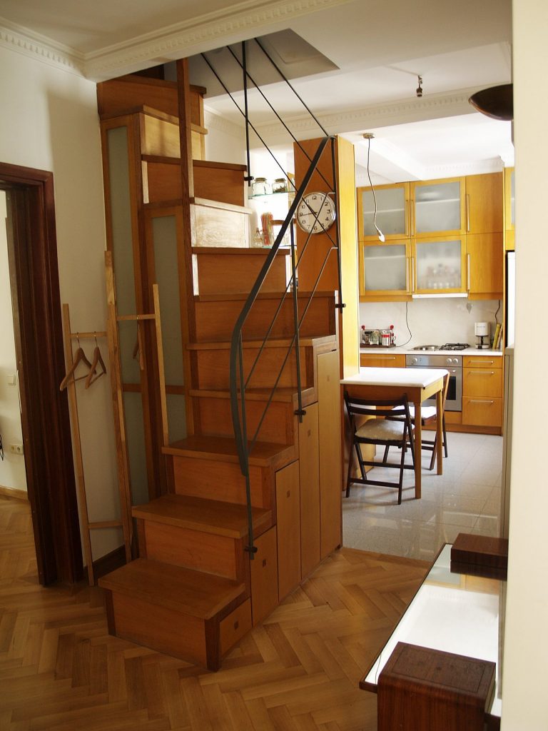 Εσωτερικό μικρής κατοικίας με ξύλινη σκάλα που οδηγεί στον επάνω όροφο και αποθηκευτικούς χώρους ενσωματωμένους κάτω από κάθε σκαλοπάτι. Δίπλα στη σκάλα υπάρχει μικρή κουζίνα με ξύλινα ντουλάπια και συσκευές, τραπεζαρία με δύο καρέκλες και δάπεδο από ξύλινο παρκέ και πλακάκια