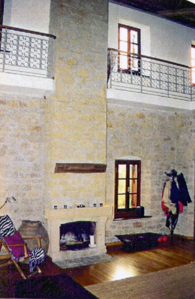 "Εσωτερικό δωματίου με ψηλό ταβάνι και πέτρινο τζάκι. Το δωμάτιο έχει ρουστίκ και άνετη ατμόσφαιρα, με πέτρινους τοίχους και ξύλινο δάπεδο. Υπάρχει μπαλκόνι με διακοσμητικά μεταλλικά κάγκελα στον επάνω όροφο και δύο παράθυρα με ξύλινα πλαίσια που επιτρέπουν το φυσικό φως να εισέλθει στο δωμάτιο