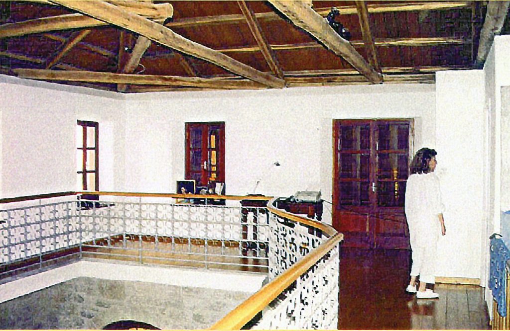 Εσωτερικό δωματίου με ξύλινη οροφή με εκτεθειμένα δοκάρια, λευκούς τοίχους και ξύλινο πάτωμα. Υπάρχει ένα άτομο που στέκεται κοντά σε τοίχο, γραφείο με υπολογιστή και γυάλινες πόρτες και παράθυρο που αφήνουν άπλετο φυσικό φως να μπαίνει