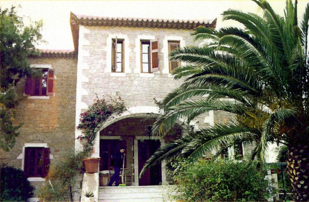 Πρόσοψη πέτρινου σπιτιού με κεραμοσκεπή και παράθυρα με ξύλινα παραθυρόφυλλα, που περιβάλλεται από πλούσια βλάστηση. Υπάρχει μια καμάρα στην είσοδο, ενώ ένα άτομο σκουπίζει τα σκαλοπάτια. Δεξιά φαίνεται ένας μεγάλος φοίνικας