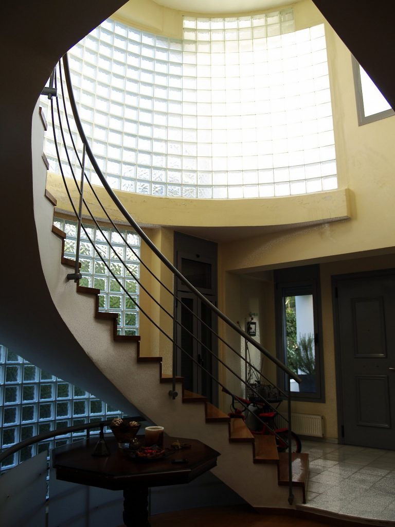 Εσωτερικό σύγχρονου σπιτιού με ξύλινη σπειροειδή σκάλα και γυάλινους τοίχους. Η σκάλα έχει ξύλινα σκαλιά και μεταλλικά κάγκελα, και οδηγεί στον επάνω όροφο. Στο κάτω μέρος της σκάλας υπάρχει ένα μικρό τραπέζι με διακοσμητικά αντικείμενα. Κοντά στην πόρτα, βρίσκεται ένα παιδικό τρίκυκλο