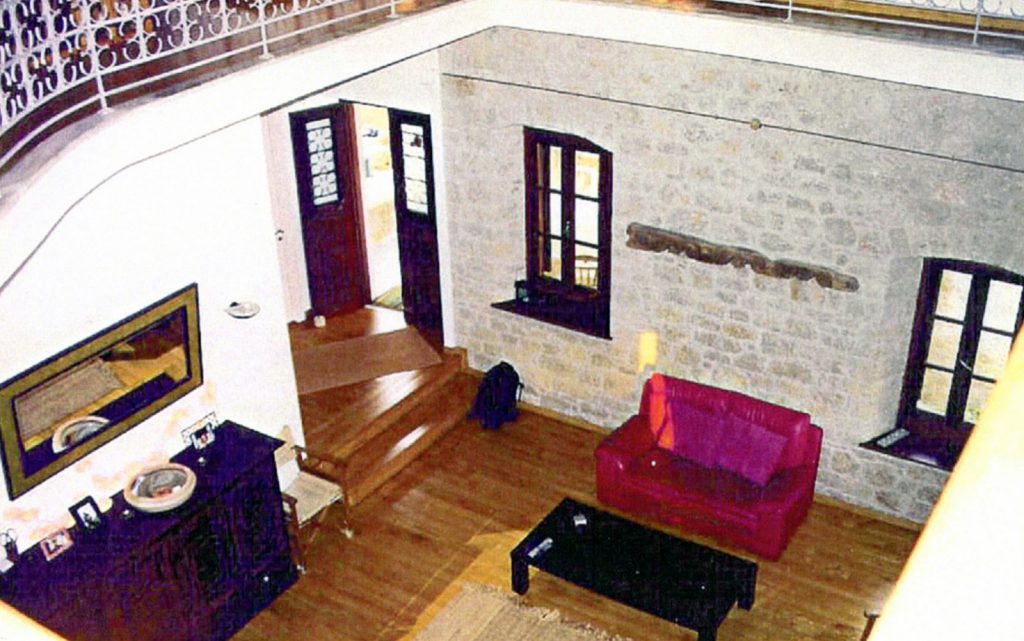 Καθιστικό με ξύλινο πάτωμα, κόκκινο καναπέ, μαύρο τραπεζάκι, τοίχο με πέτρα και παράθυρα, σκάλες που οδηγούν σε πόρτα, μαύρο ντουλάπι με καθρέπτη και διακοσμητικά κάγκελα