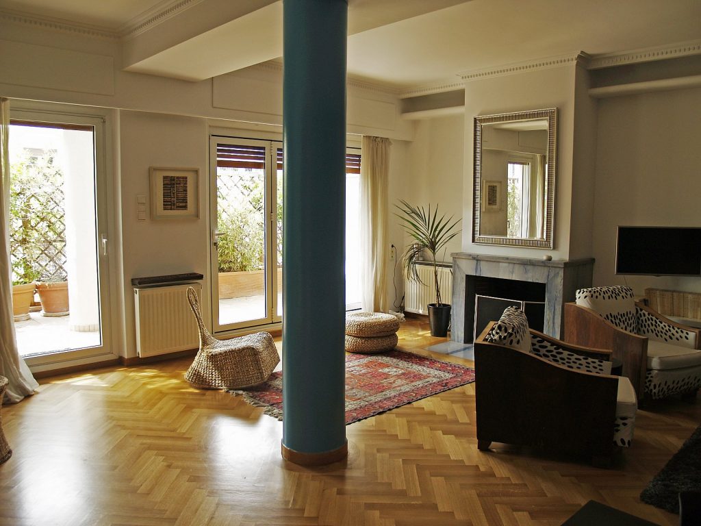 Φωτεινό σαλόνι με μοντέρνο και άνετο σχεδιασμό, μεγάλο μπλε κολώνα στο κέντρο, τζάκι με μεγάλο καθρέπτη πάνω του, σύγχρονα και ρουστίκ έπιπλα, δύο καρέκλες από λυγαριά, μοκέτα με μοτίβο και καρέκλες με μαύρο-άσπρο σχέδιο. Υπάρχουν ξύλινο παρκέ δάπεδο και μεγάλες γυάλινες πόρτες που ανοίγουν σε μπαλκόνι με γλάστρες