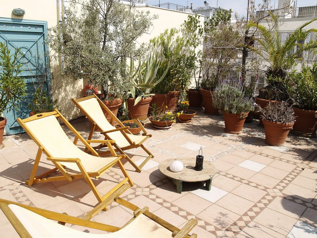 Βεράντα με ξύλινα ξαπλώστρες και τραπέζι με φυτά και φανάρια, περιτριγυρισμένη από δέντρα και πρασινάδα. Φόντο με κτίρια και κίτρινο τοίχο με μπλε πόρτα