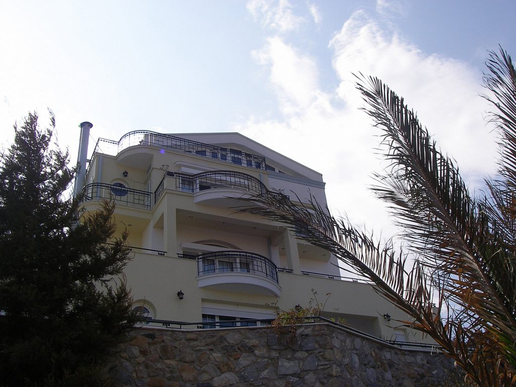 Πολυώροφο κτίριο με μπαλκόνια και κιγκλιδώματα, περιτριγυρισμένο από δέντρα και φυτά, με καθαρό ουρανό στο παρασκήνιο
