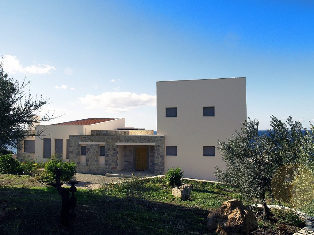 Μοντέρνο σπίτι με μοναδικό αρχιτεκτονικό σχεδιασμό, συνδυασμό πέτρινης και λείας επιχρισμένης πρόσοψης, επίπεδη στέγη και ορθογώνια παράθυρα. Μπροστινή είσοδος με πέτρινη πρόσοψη και ξύλινη πόρτα. Πράσινο περιβάλλον, θάλασσα στο βάθος και γαλάζιος ουρανός