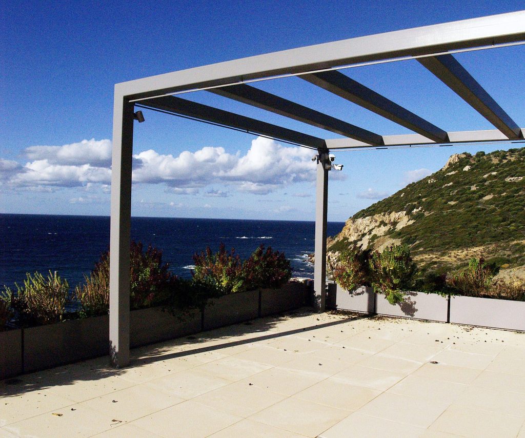 Βεράντα με μοντέρνα πέργκολα που βλέπει στη θάλασσα, γκρι δοκοί και κολώνες, πλακίδια ανοιχτού χρώματος και φυτά στην άκρη. Υπέροχη θέα στον ωκεανό και λοφώδη πράσινη περιοχή.