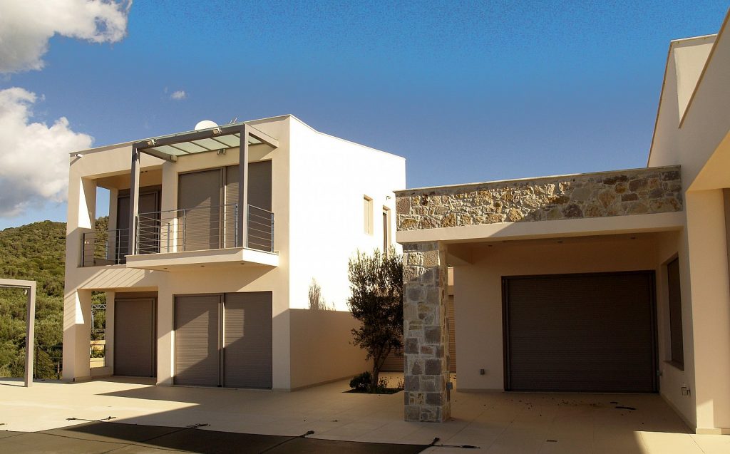 Διώροφο μοντέρνο σπίτι με επίπεδη στέγη και μεγάλα παράθυρα με ρολά, μπαλκόνι με γυάλινα και μεταλλικά κάγκελα, και πέτρινη πρόσοψη. Το σπίτι βρίσκεται σε προαστιακή περιοχή με λόφους και πράσινο στο βάθος