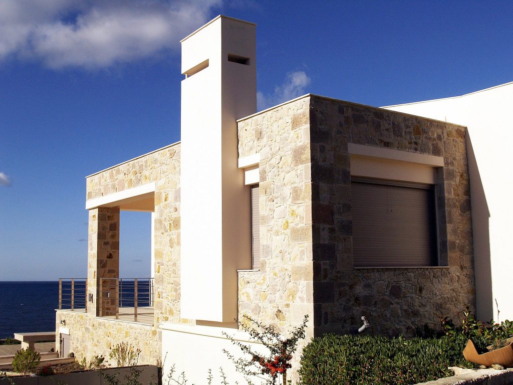 Μοντέρνο σπίτι με πέτρινη πρόσοψη και λευκή καμινάδα, μεγάλο παράθυρο με κλειστό παντζούρι, και μπαλκόνι με μεταλλικό κάγκελο που βλέπει στη θάλασσα